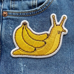 May include: Embroidered patch featuring a yellow banana slug design. The slug's body is a bright yellow, with a brown outline and a white border. The patch is sewn onto a blue denim fabric.