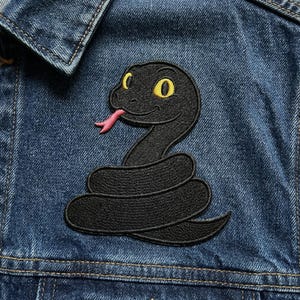 May include: A black embroidered snake patch with yellow eyes and a pink tongue is sewn onto a blue denim jacket. The snake is coiled and has a friendly expression. The jacket has a classic collar and visible stitching.