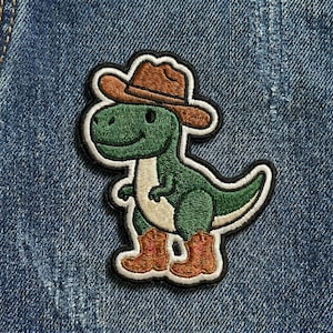 May include: Embroidered patch featuring a green dinosaur wearing a brown cowboy hat and boots. The dinosaur has a white belly and is outlined in black. The patch is on a blue denim background.