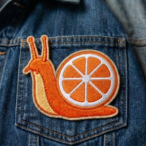 May include: An embroidered patch featuring an orange snail with a slice of orange for a shell. The patch is primarily orange with white and yellow accents. It is attached to a blue denim jacket pocket.