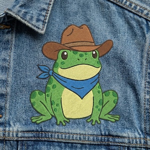 May include: Embroidered patch featuring a green frog wearing a brown cowboy hat and a blue bandana. The frog has yellow markings and is set against a denim jacket.