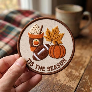 May include: A circular embroidered patch with a cream-colored background and a dark brown border. The design features a coffee cup with whipped cream, a football, a maple leaf, and a pumpkin. The text TIS THE SEASON is embroidered in brown.