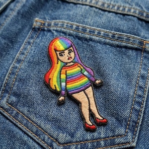 May include: Embroidered patch featuring a cartoon figure with rainbow hair and a rainbow striped top. The figure is wearing a short skirt and red shoes. The patch is set against a blue denim background.