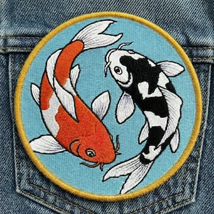 May include: Embroidered patch featuring two koi fish in a circular design. One fish is orange and white, the other is black and white, set against a light blue background with a gold border. The patch is sewn onto a denim fabric.