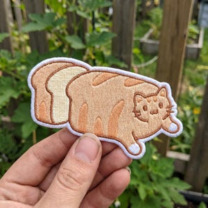 May include: Embroidered patch featuring a cartoon cat shaped like a slice of bread. The cat is tan with darker brown stripes and white paws. The bread slice is light brown with a white border.