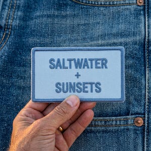 May include: A rectangular light blue embroidered patch with a dark blue border. The text SALTWATER + SUNSETS is stitched in dark blue capital letters in the center of the patch. The patch is positioned against a blue denim fabric background.