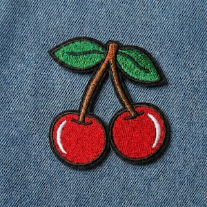 May include: Embroidered cherry patch featuring two red cherries with white highlights, green leaves, and a brown stem. The patch is outlined in black and set against a blue denim background.