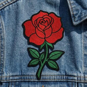 May include: A red rose patch with green leaves and stem is sewn onto a denim jacket. The rose is embroidered with a black outline, adding definition to the petals. The denim jacket is a light blue wash.