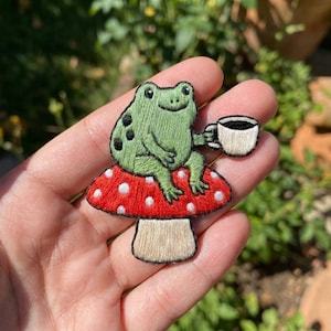May include: An embroidered patch featuring a light green frog with black spots sitting on a red mushroom with white polka dots. The frog is holding a small white cup with a black rim. The mushroom has a tan stem and a rounded red cap.