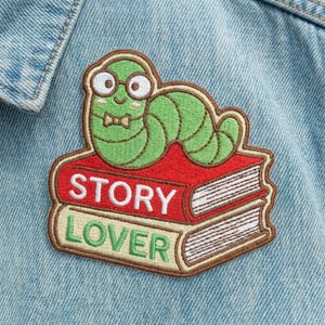 May include: Embroidered patch featuring a green worm wearing glasses and a bow tie, perched atop two books. The top book is red with "STORY" in white, and the bottom book says "LOVER" in green.