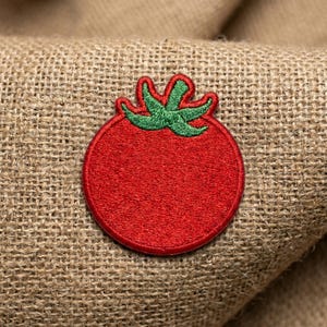 May include: A red embroidered tomato patch with green leaves. The patch is circular and has a textured appearance, set against a burlap background. This patch is suitable for clothing or accessories.