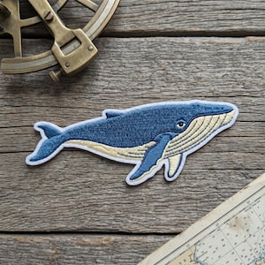 May include: Embroidered patch featuring a blue whale with a cream-colored belly and accents. The whale is set against a white border. The patch is on a wooden surface, with a nautical instrument and a map in the background.
