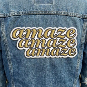 May include: A blue denim jacket with a large embroidered patch on the back. The patch features the word "amaze" repeated three times in a gold script font with a white border.
