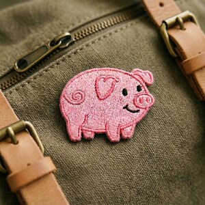 May include: A pink embroidered pig patch with a heart-shaped ear and a curly tail. The patch is sewn onto a green canvas material, with leather straps and a zipper visible in the background.