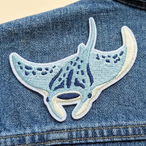 May include: Embroidered patch featuring a light blue manta ray design with darker blue and cream accents. The patch is sewn onto a denim jacket, showcasing the intricate details of the sea creature.