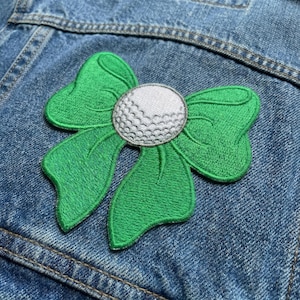 May include: Embroidered green bow patch with a white golf ball center, set against a denim background. The patch features detailed stitching and a playful design, perfect for adding a unique touch to clothing or accessories.