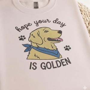 May include: A light pink sweatshirt with the embroidered phrase "hope your day is GOLDEN." The design features a cartoon golden retriever wearing a blue bandana, with paw prints on either side.