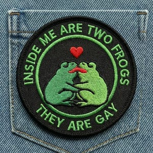 May include: Round black patch with green text and a design of two kissing green frogs. The text reads "INSIDE ME ARE TWO FROGS THEY ARE GAY" with a red heart above the frogs. The patch is on a denim background.