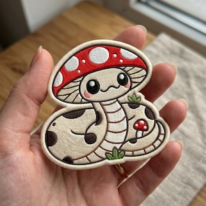 May include: An embroidered patch featuring a cartoon snake with a mushroom cap head. The snake has a cream-colored body with brown spots, a red mushroom cap with white spots, and a small red mushroom beside it, all outlined in cream thread.