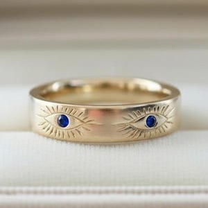 May include: A gold ring featuring an engraved eye pattern and two small blue gemstones. The ring is presented in a beige jewellery box with a soft, velvety lining. The ring has a wide, flat band.