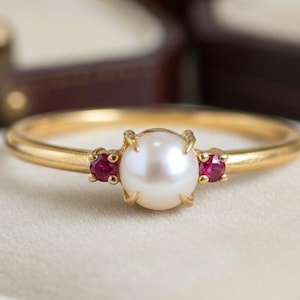 Könnte beinhalten: Ein goldener Ring mit einer zentralen weißen Perle und zwei kleineren roten Edelsteinen auf beiden Seiten. Der Ring liegt auf einer weißen Oberfläche, mit einem verschwommenen Hintergrund aus offenen Schmuckkästchen.