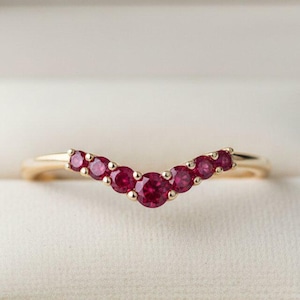 May include: A gold ring with a curved design, set with seven round, vibrant red gemstones. The ring is displayed inside a light pink jewelry box, showcasing its elegant and delicate appearance. The gemstones are evenly spaced along the curve.