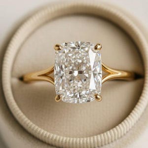 May include: A radiant-cut diamond engagement ring with a gold band, set in a beige velvet ring box. The diamond is rectangular with cut corners, and the gold band has a delicate, split-shank design.