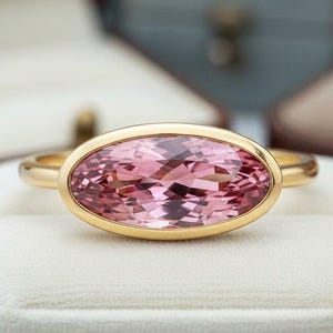 Op de afbeelding: Een gouden ring met een ovale, roze edelsteen. De edelsteen is gezet in een gouden bezel. De ring wordt gepresenteerd op een witte fluwelen ondergrond. Het ontwerp van de ring is eenvoudig en elegant. Op de achtergrond staan open sieradendoosjes.