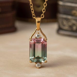 May include: A gold necklace with a rectangular pendant featuring a bi-colour gemstone. The gemstone transitions from pink to green, with small clear stones on the pendant. The necklace is displayed against a blurred background.