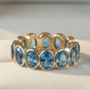 Peut inclure: Une bague en or ornée d'une série de pierres précieuses ovales bleu clair. La bague est sertie dans un anneau en or et est présentée dans un écrin blanc. Les pierres sont espacées uniformément.
