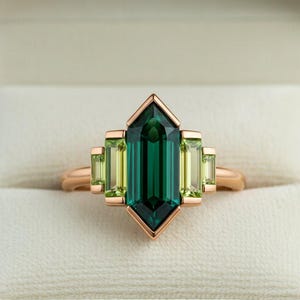 May include: A rose gold ring featuring a large, emerald-cut, deep green gemstone at the center, flanked by smaller, rectangular, light green stones. The ring is set in a geometric design, presented in a white jewelry box.