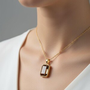May include: A gold-colored necklace with a rectangular, brown gemstone pendant. The pendant is set in a gold frame and hangs from a delicate gold chain. The necklace is worn against a white top.
