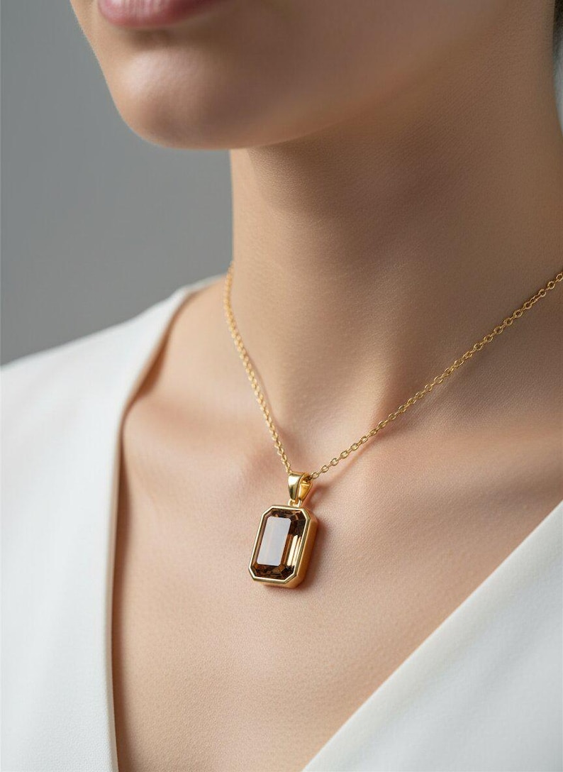May include: A gold-colored necklace with a rectangular, brown gemstone pendant. The pendant is set in a gold frame and hangs from a delicate gold chain. The necklace is worn against a white top.