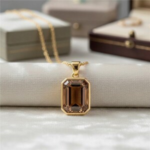 May include: A gold-colored necklace with a rectangular, brown gemstone pendant. The pendant is set in a gold bezel and hangs from a delicate gold chain. The necklace is displayed on a white, textured surface.