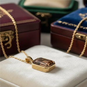 May include: A gold necklace with a rectangular, brown gemstone pendant. The pendant is set in a gold bezel and hangs on a delicate gold chain. The necklace is displayed on a white surface, with jewelry boxes in the background.