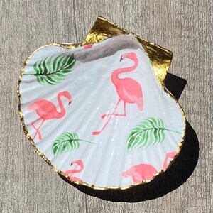 May include: A decorative seashell with a gold-painted edge. The interior features a white background with pink flamingo illustrations and green leaf accents. The seashell is resting on a wooden surface.