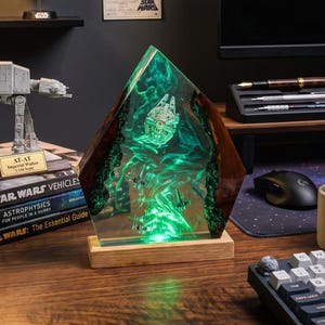 Peut inclure: Un bureau sur le thème de Star Wars avec une sculpture en résine verte lumineuse représentant le Faucon Millenium. D'autres éléments incluent un modèle AT-AT, des livres Star Wars, une affiche et un cactus en pot.