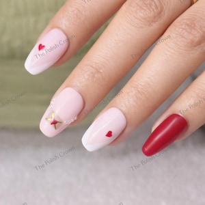 May include: A hand with manicured nails in various designs. Some nails are light pink with white tips and red heart accents. One nail is a solid deep red. A gold ring with a clear stone is also visible.