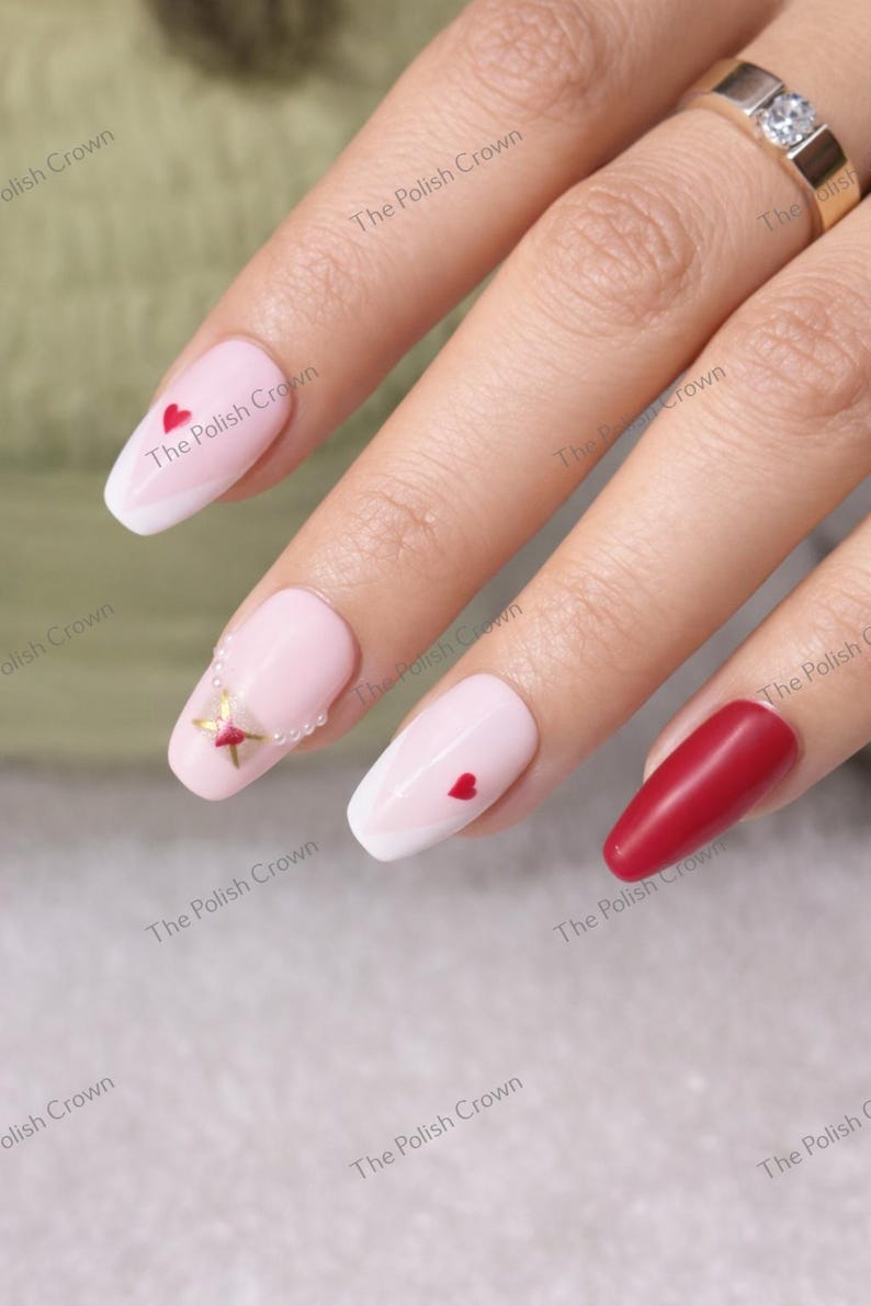 May include: A hand with manicured nails in various designs. Some nails are light pink with white tips and red heart accents. One nail is a solid deep red. A gold ring with a clear stone is also visible.