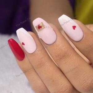 May include: A hand with manicured nails. The nails are painted in various designs, including solid red, white tips, pink with a red heart, and pink with a gold and red floral design and pearls. The nails are oval-shaped.