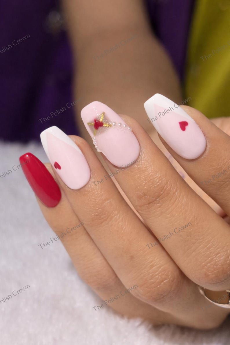 May include: A hand with manicured nails. The nails are painted in various designs, including solid red, white tips, pink with a red heart, and pink with a gold and red floral design and pearls. The nails are oval-shaped.