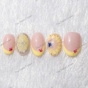 Puede incluir: Un conjunto de cinco uñas artificiales con base rosa claro y puntas amarillas. Cada uña presenta diseños únicos, incluyendo acentos florales, un patrón de estrella y adornos dorados. El texto "The Polish Crown" es visible.