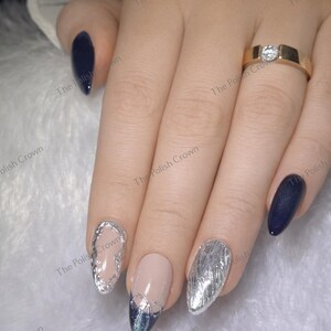 May include: Close-up of a hand with manicured nails. Some nails are painted navy blue, while others feature silver designs and a nude base. A gold ring with a diamond is on the ring finger. The Polish Crown is printed on the image.