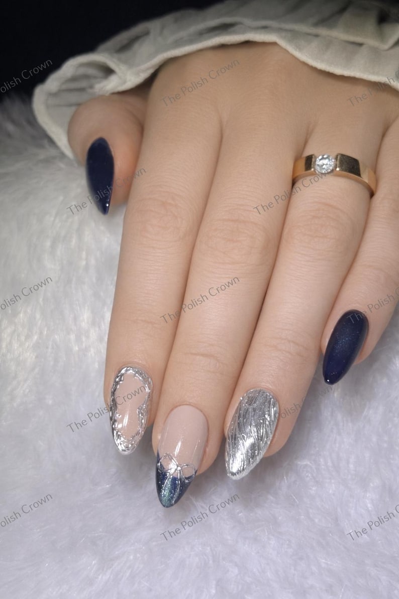 May include: Close-up of a hand with manicured nails. Some nails are painted navy blue, while others feature silver designs and a nude base. A gold ring with a diamond is on the ring finger. The Polish Crown is printed on the image.