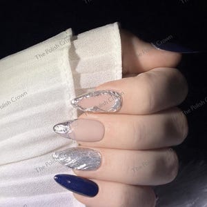 May include: Close-up of a hand with manicured nails. The nails feature a variety of designs, including silver foil accents, a nude matte finish, and a deep navy blue polish. The text "The Polish Crown" is visible in the background.