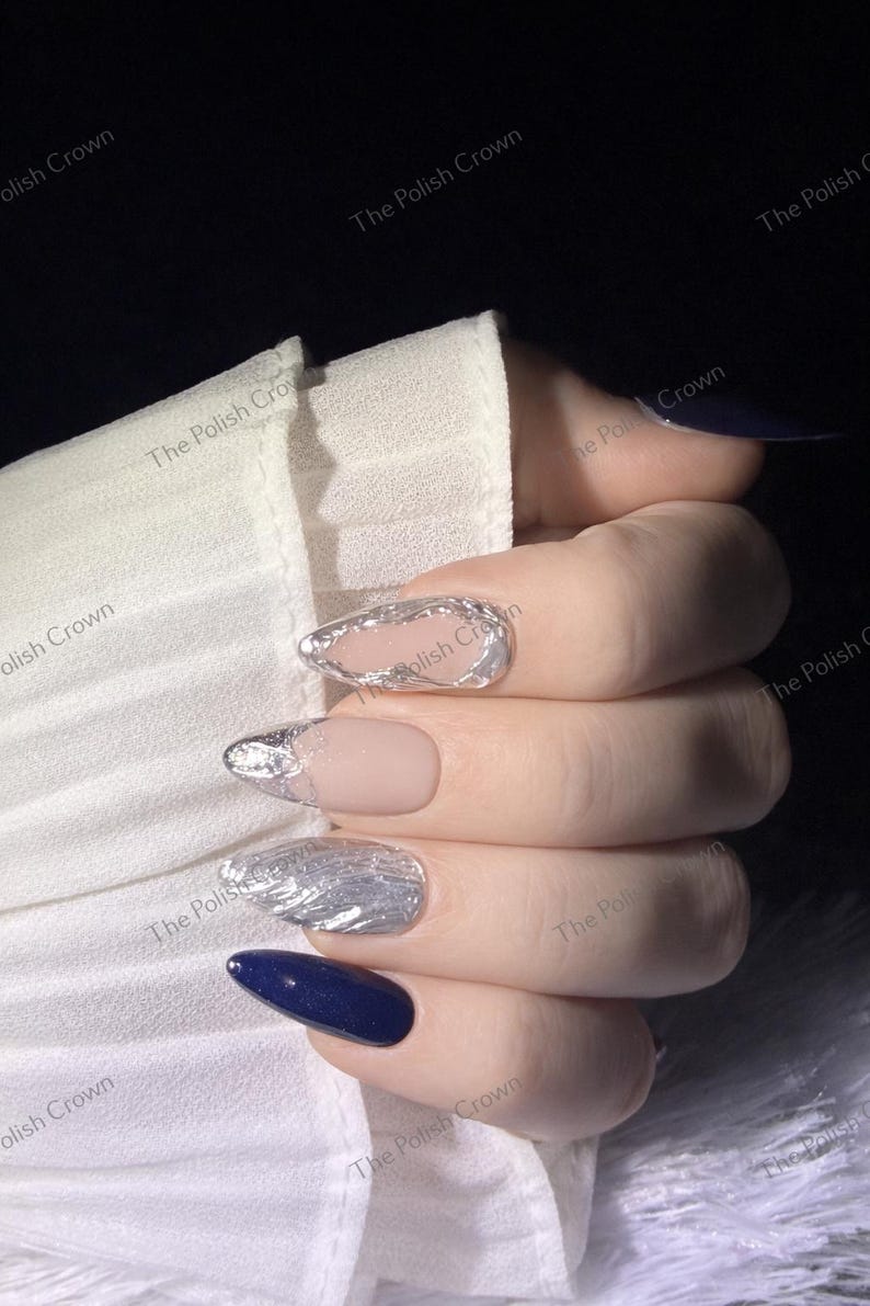 May include: Close-up of a hand with manicured nails. The nails feature a variety of designs, including silver foil accents, a nude matte finish, and a deep navy blue polish. The text "The Polish Crown" is visible in the background.