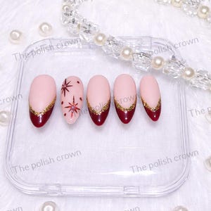 May include: A set of five artificial nails with a light pink base, dark red tips, and gold glitter accents. One nail features a red starburst design. The nails are displayed in a clear plastic case, with a pearl necklace in the background.