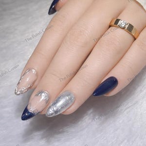 May include: Close-up of a hand with long, pointed nails. Some nails are painted navy blue, others are silver with intricate designs. A gold ring with a single diamond is on the ring finger. The Polish Crown is printed on the image.