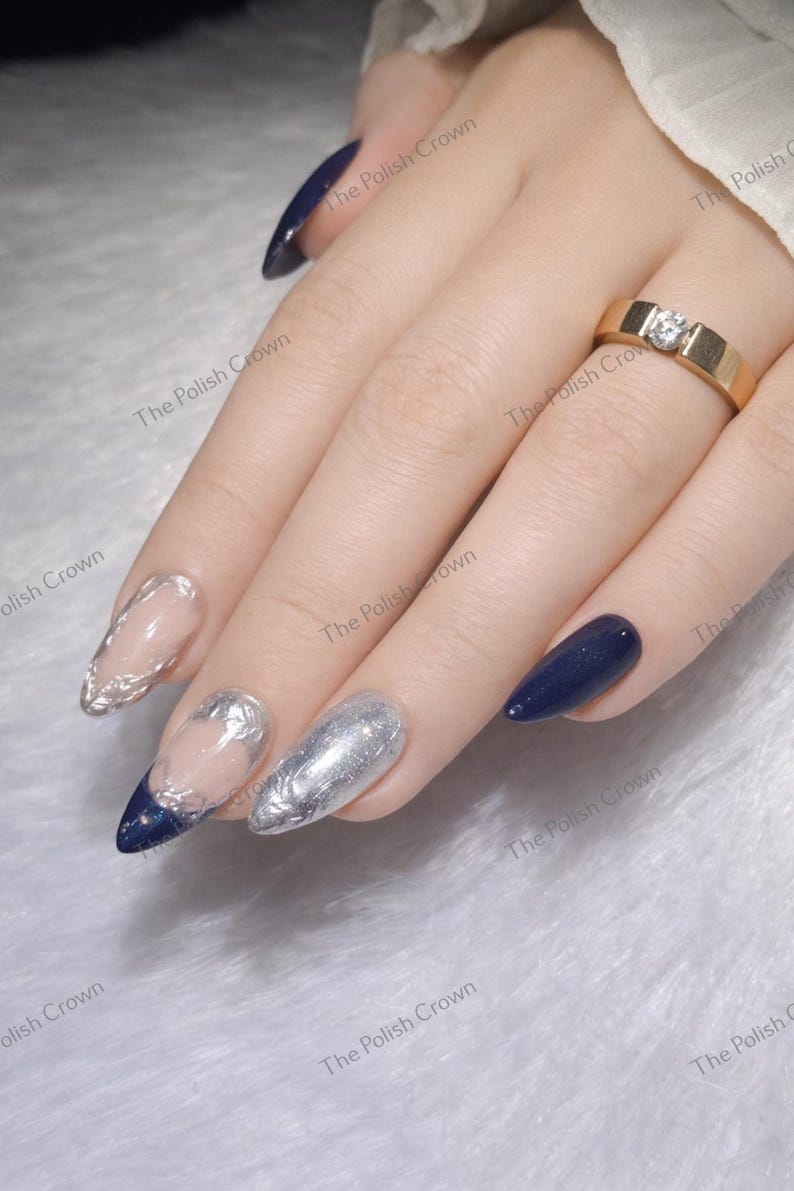 May include: Close-up of a hand with long, pointed nails. Some nails are painted navy blue, others are silver with intricate designs. A gold ring with a single diamond is on the ring finger. The Polish Crown is printed on the image.