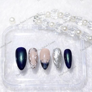 May include: A set of five artificial nails in a clear case. The nails feature various designs, including a dark blue with green shimmer, a silver textured nail, and a nude nail with silver accents. The text "The Polish Crown" is visible.
